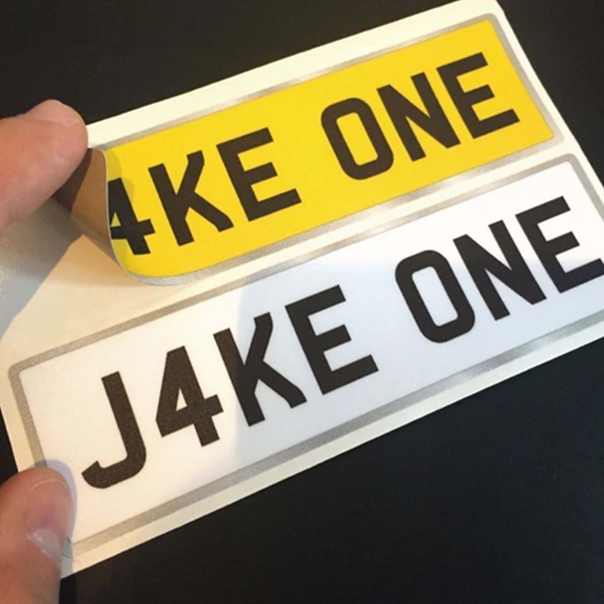 A person holding a personalized "Jake One" registration plate sticker for kids cars from Toy's Number Plates store.